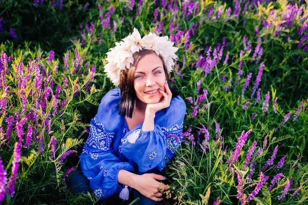 Giovane bella donna con corona di fiori seduta in campo viola. Ritratto di ragazza in abiti blu . — Foto Stock