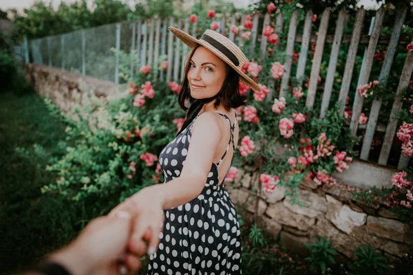 Elegante jonge vrouw in retro stijl jurk en hoed houden hand van haar man op rozen achtergrond. Volg mij naar. Travel concept. — Stockfoto