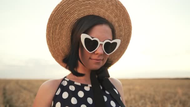 Retrato de mujer con estilo en el campo de trigo. Chica a la antigua en sombrero de paja, gafas en forma de corazón, vestido de lunares negro. Viajes, moda, concepto de naturaleza — Vídeo de stock