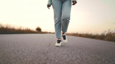 Kot pantolon ve beyaz ayakkabı yabancı kız kamera ileri asfalt pürüzsüz yolda yürüyor. Modern spor ayakkabı kavramı, seyahat, yaz doğada yürüyüş.