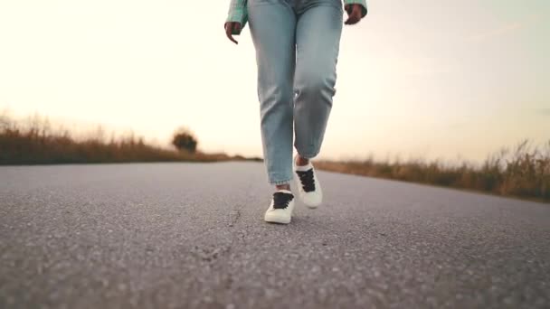 Menina mais estranha em jeans e sapatos brancos está andando ao longo do asfalto estrada lisa para a frente para camera.Concept de tênis modernos, viagens, caminhadas na natureza verão . — Vídeo de Stock