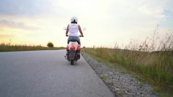 Vrouw rijdt op retro scooter langs de snelweg in de zomer bij zonsondergang. Prachtige Italiaanse natuur. Camera schiet van achteren. — Stockvideo