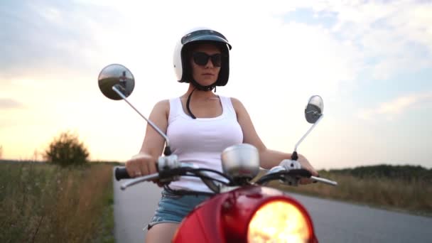 Junges hübsches Frauenporträt. Mädchen mit Helm und Brille sitzt auf rotem Retro-Roller vor Naturkulisse. — Stockvideo