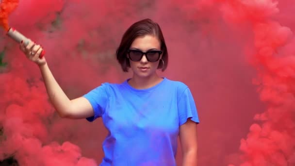 劇的な気分の照明で赤い煙の爆弾や手榴弾を持つ若いかわいい女性。青いTシャツとサングラスを着た女の子が丸い動きをする. — ストック動画