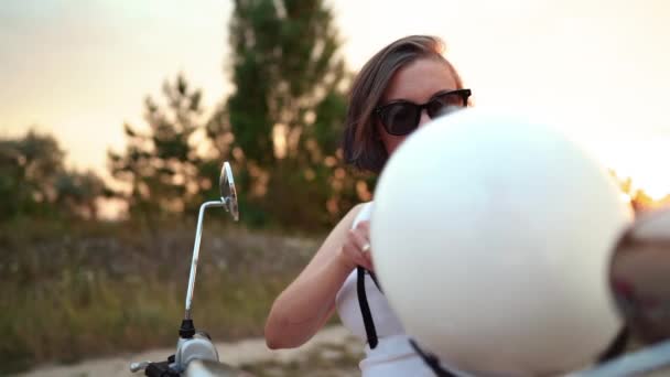 Mooie vrouw zet motorfiets helm terwijl zittend op vintage rode scooter op de snelweg in de zomer bij zonsondergang. — Stockvideo