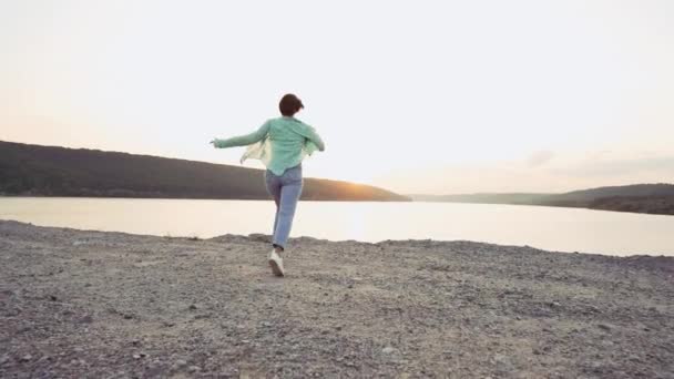 Mujer joven corre al acantilado por encima de ancho río o lago con montañas. Concepto de libertad, viajes, éxito, logro de metas — Vídeo de stock