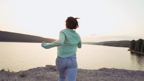 Mujer joven corre al acantilado por encima de ancho río o lago con montañas. Concepto de libertad, viajes, éxito, logro de metas — Vídeo de stock