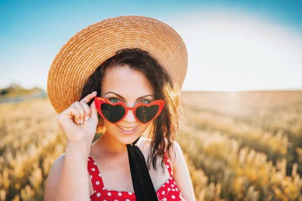 Portret stylowej kobiety w dziedzinie pszenicy. Staromodny dziewczyna w słomkowym kapeluszu, okulary w kształcie serca, czerwona polka dot sukienka i szminka. Podróże, moda, koncepcja przyrody — Zdjęcie stockowe