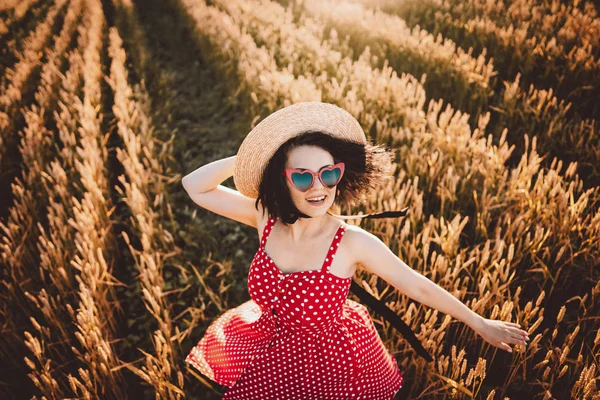 Donna nel campo di grano. Ragazza all'antica in cappello di paglia, occhiali a forma di cuore, abito rosso a pois. Viaggio, moda, natura — Foto Stock