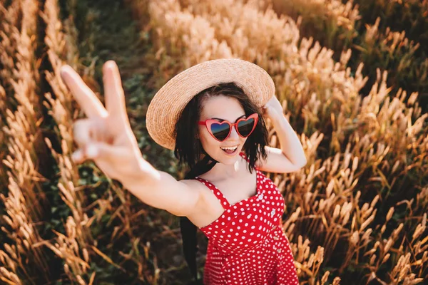 Жінка в пшеничному полі, що показує знак ОК. Стара модна дівчина в солом'яному капелюсі, окуляри у формі серця, червона сукня-полька. Подорожі, мода, концепція природи — стокове фото
