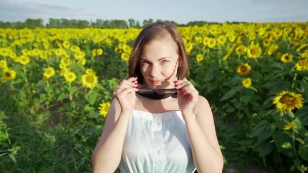 Jonge mooie vrouw zet op zonnebril in sunflowersop zoek naar camera en glimlachend — Stockvideo