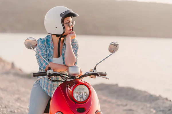 Young pretty womans portrait. Girl in white helmet and eyeglasses sitting on red retro scooter at nature background. — Stock Photo, Image