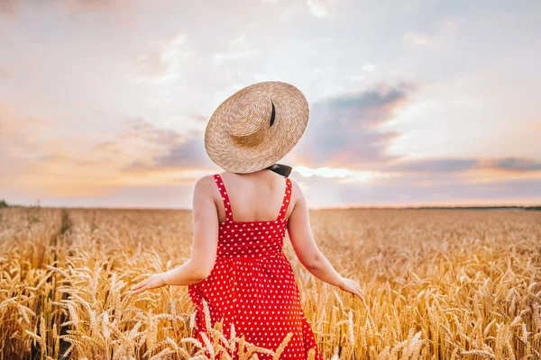 Unrecognizable woman in red dress with straw hat standing in field. Golden hour. Harvest, travel concept.