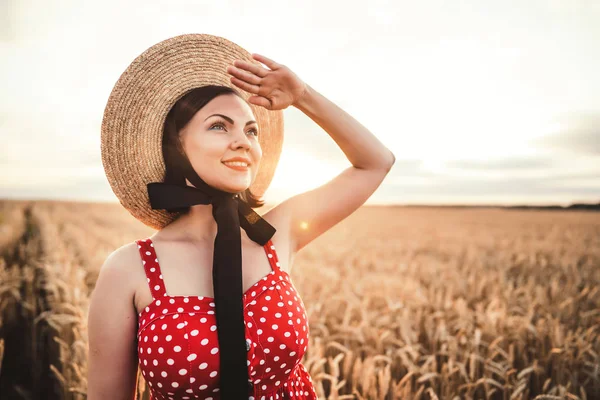 Donna allegra in abito rosso e cappello sorridente e guardando in lontananza nascondendo mano dal sole nel campo di grano dorato. Bellezza, natura, viaggio, raccolto, armonia concetto . — Foto Stock