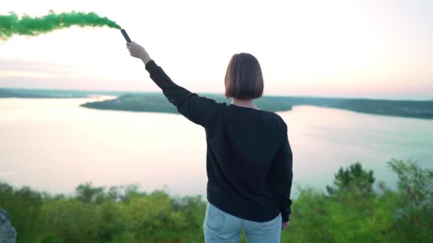 Chica de negro haciendo movimientos redondos de pie en la roca sobre el río. Mujer joven con bomba de humo verde o granada en el fondo de la naturaleza dramática — Vídeos de Stock