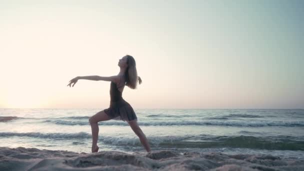 Jeune jolie ballerine en robe noire dansant ballet sur la mer ou la plage de sable de l'océan à la lumière du matin. Concept d'étirement, art, beauté de la nature — Video