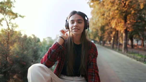 Ung tonåring lyssnar på musik via hörlurar i parken.Flicka i röd rutig skjorta ler, dansar till rytm.Begreppet studentliv, frihet, modern ungdom — Stockvideo
