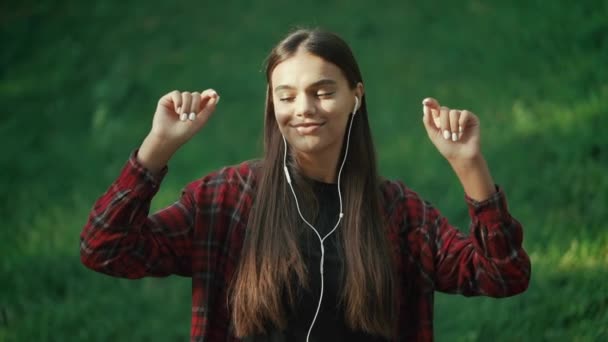 Retrato de menina atraente dançando com fones de ouvido no parque. Mulher a sorrir. move-se para o ritmo.Aparência amigável do moderno hipster da moda — Vídeo de Stock