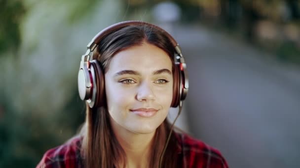 Close-up van een meisjes gezicht in een koptelefoon. Een jonge vrouw geniet van muziek terwijl ze in een park zit — Stockvideo