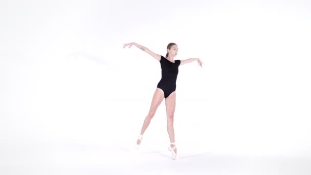 Балерина в черном костюме репетирует в балете в светлой студии. Молодая девушка, изолированная на белой стене, танцует классический танец в пуантах. Медленное движение . — стоковое видео