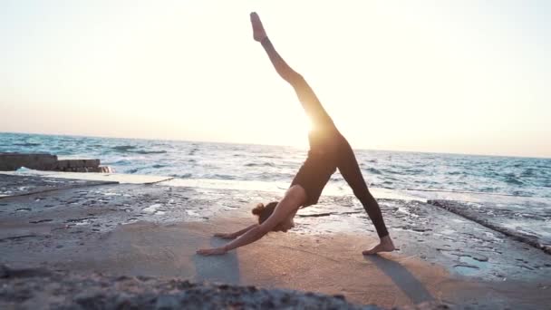 Mujer delgada en negro practicando yoga cerca del mar o el océano durante la luz del amanecer. Flexibilidad, estiramiento, fitness, estilo de vida saludable — Vídeos de Stock