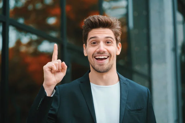 Portrait of young thinking pondering businessman having idea moment pointing finger up on office building background. Smiling happy student man showing eureka gesture. — Stock Photo, Image