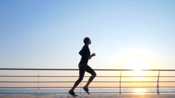 Sportliches junges Mädchen joggt morgens am Holzdamm des Meeres. Silhouette eines Mädchens im Sportkostüm. schönes Sonnenlicht. Konzept eines gesunden Lebensstils. — Stockvideo