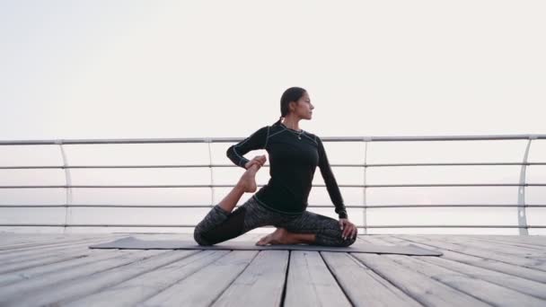 Giovane donna bruna che fa esercizi di yoga su argine di legno al mattino. Ragazza in costume sportivo nero. Allungamento, pratica, concetto di stile di vita sano — Video Stock