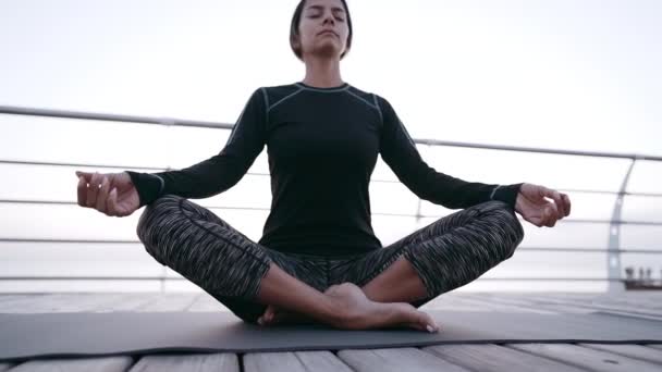 Konzentriertes Mädchen in Schwarz praktiziert Yoga-Meditation an der hölzernen Strandpromenade. Frau in Lotuspose. Dehnen, üben, gesundes Lebensstilkonzept — Stockvideo