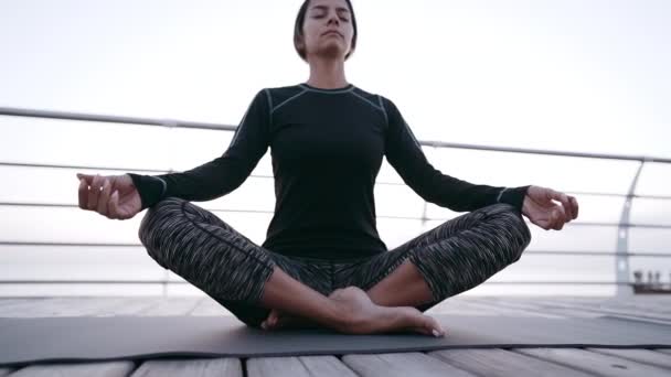 Concentrated girl in black practicing in yoga meditation on wooden seafront. Woman in lotus pose. Stretching, practice, healthy lifestyle concept — Stock Video