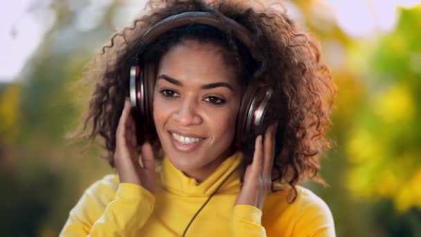 Zbliżenie dziewcząt twarz w słuchawkach. Młoda kobieta w żółtym lubi muzykę siedząc w parku. Zwolnionym. — Wideo stockowe