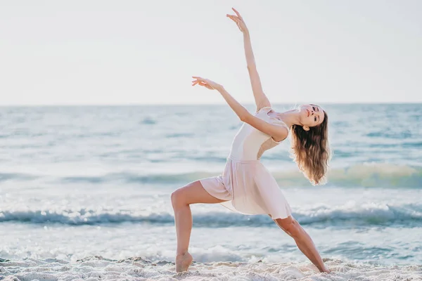 Joven bailarina bonita en vestido beige bailando ballet en el mar o playa de arena del océano en la luz de la mañana. Concepto de estiramiento, arte, belleza de la naturaleza — Foto de Stock