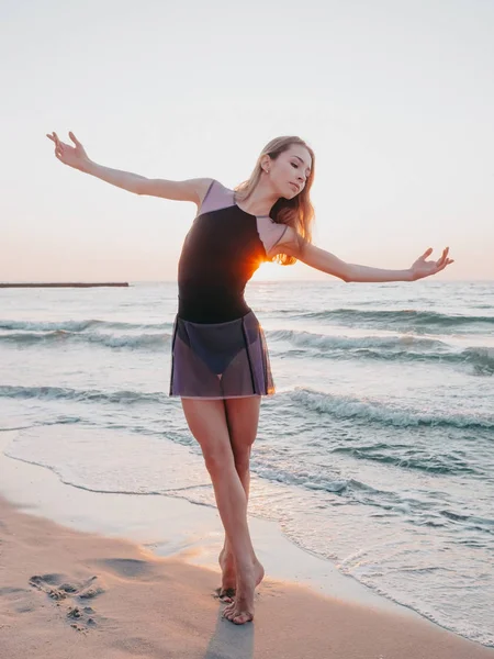 Танцующая балерина в черном шелковом платье на набережной возле океана или моря на восходе или закате солнца. Силуэт молодой женщины с длинными волосами, практикующей классические упражнения с эмоциями — стоковое фото