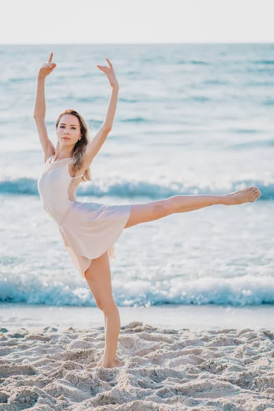 Jonge mooie ballerina in beige jurk dansen ballet op zee of Oceaan zandstrand in ochtend licht. Concept van stretching, kunst, natuur schoonheid — Stockfoto