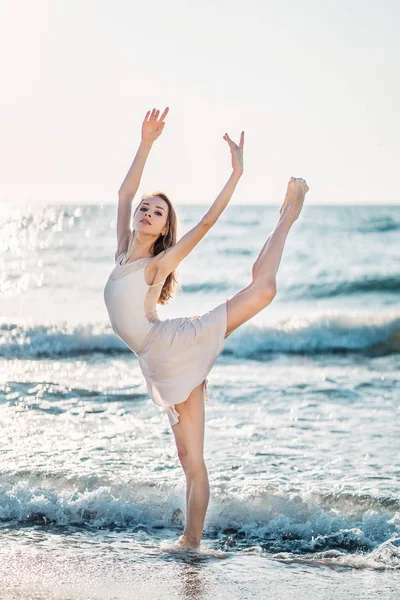 Jonge mooie ballerina in beige jurk dansen ballet op zee of Oceaan zandstrand in ochtend licht. Concept van stretching, kunst, natuur schoonheid — Stockfoto