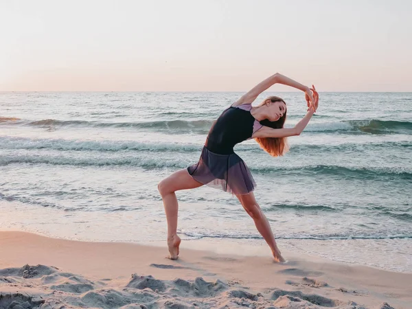 Bailarina bonito flexível em vestido preto dança balé na praia de areia do mar na luz da manhã. Conceito de alongamento, arte, beleza da natureza — Fotografia de Stock