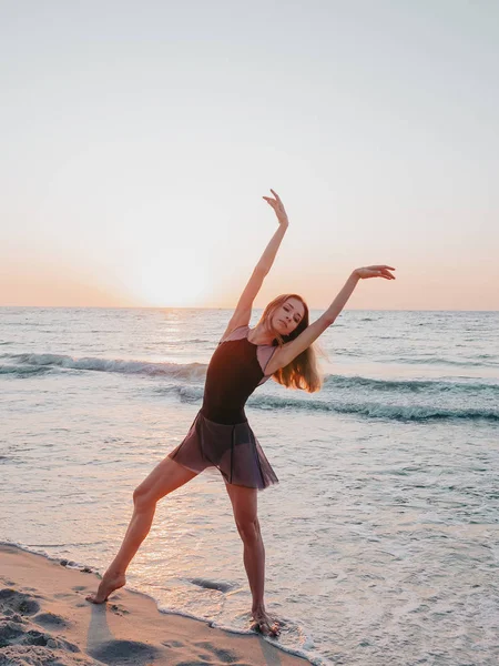 Bailarina bonito flexível em vestido preto dança balé na praia de areia do mar na luz da manhã. Conceito de alongamento, arte, beleza da natureza — Fotografia de Stock