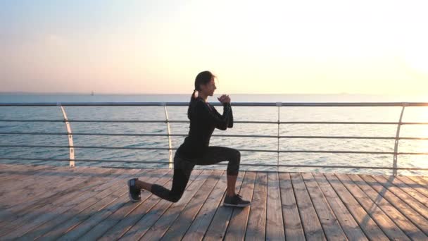 Chica atlética joven realiza sentadillas en terraplén de madera junto al mar en la mañana temprano. Estilo de vida saludable, entrenamiento, concepto de entrenamiento . — Vídeos de Stock