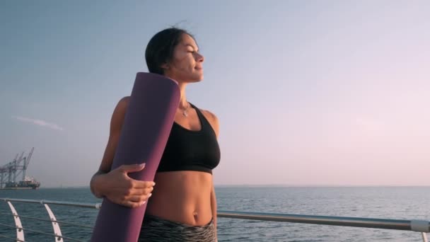 年轻性感的女运动员散步做晨练。 拿着瑜伽垫的女孩 海或海的背景。 夏日。 慢动作. — 图库视频影像