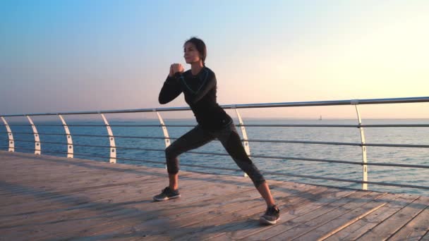 Young athletic girl performs squats on wooden embankment by the sea in early morning. Healthy lifestyle, coaching, training concept. — Stock Video