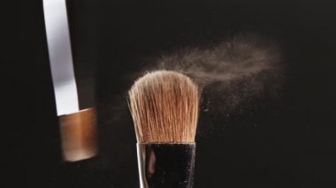 İki makyaj fırçası birbirine siyah arka plan ve kozmetik parçacıkları üzerinde dokunur. Gölgeler birbirinden ayrılır, patlamanın etkisi. Güzellik endüstrisi, enstrümanlar, yavaş çekim.