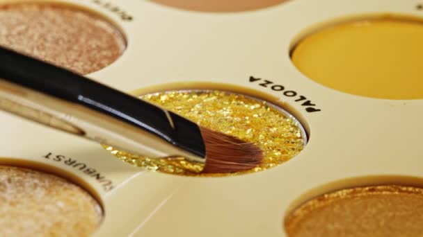 Sikat memperoleh glitter kuning dari isi ulang pada tumpukan yang akan digunakan dalam make-up. Seniman yang bekerja dengan eyeshadows palet. Rincian proses kerja, alat-alat dalam industri kecantikan. Kosmetik dekoratif. — Stok Video
