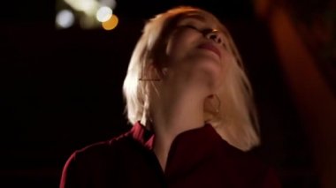 Çekici bir kadının portresi saçlara dokunur, dalgalar saçı dalgalanır ve koyu arkaplanda sarı saç stiliyle mutluluğa ulaşır. Gülümseyen güzel çekici kız. Geceleri şehir. Burun delici. Yavaş çekim.