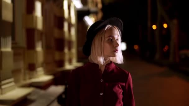 Hipster mulher loira de chapéu andando na rua da cidade noite. Rapariga na cidade europeia sozinha. Arquitetura iluminada com luz. Movimento lento . — Vídeo de Stock