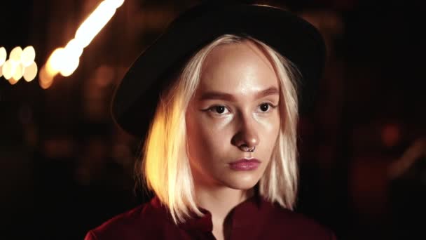 Porträtt av trendiga hipster kvinna med blond frisyr står på stadens gata på natten. Hatt, näsa piercing. Mystisk flicka. Långsamma rörelser. — Stockvideo
