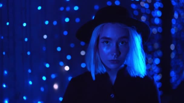 Tisíciletá záhadná hezká žena s blonďatým účesem kráčející v noci poblíž zářící neonové stěny. Modré vlasy, hipsterský klobouk, piercing v nose. Záhadná dívka. Zpomalený pohyb. — Stock video