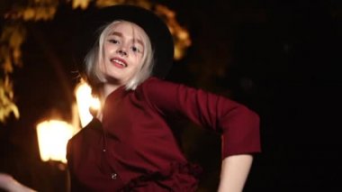 Millennial hippi kadın, sarışın saç stili ile gecenin karanlığında sokak fenerinin altında dans ediyor. Şapka, burun piercing 'i. Komik kız. Yavaş çekim.