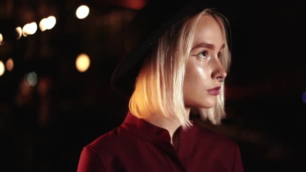 Retrato de mujer hipster de moda con peinado rubio de pie en la calle de la ciudad por la noche. Sombrero, piercing en la nariz. Una chica misteriosa. Movimiento lento . — Vídeo de stock