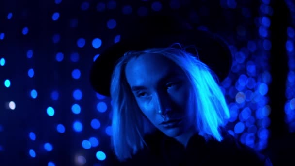 Millennial enigmática bonita menina loira penteado perto brilhante parede de néon à noite. Cabelo azul, chapéu hipster, piercing no nariz. Mulher misteriosa. Movimento lento . — Vídeo de Stock