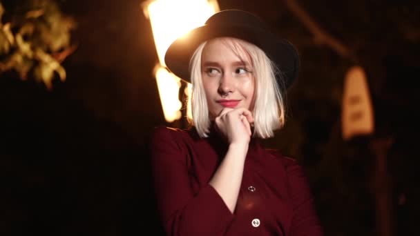 Portret van millennial hipster vrouw met blond haar onder de lantaarn van de stad straat 's nachts. Hoed, neuspiercing. Grappig meisje. Langzame beweging. — Stockvideo